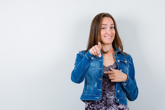 Portrait of young woman pointing at front in denim jacket, dress and looking jovial front view