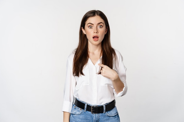 Portrait of young woman pointing at herself with disbelief, surprised face expression, being named or chosen, standing over white background