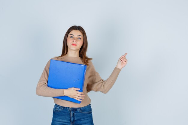 Portrait of young woman pointing up, keeping folder in sweater, jeans and looking thoughtful, front view .