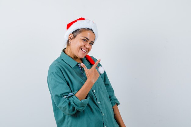 Portrait of young woman pointing at upper right corner in shirt, Santa hat and looking joyful front view