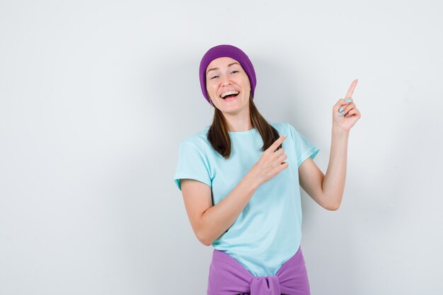 Portrait of young woman pointing at upper right corner in t-shirt, beanie and looking merry front view