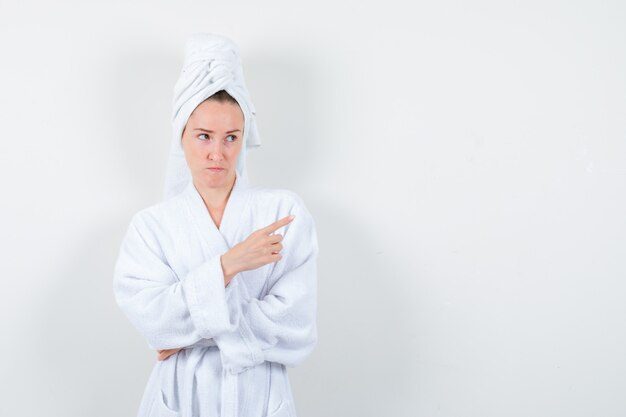 Portrait of young woman pointing at upper right corner in white bathrobe, towel and looking hesitant front view
