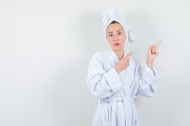 Portrait of young woman pointing at upper right corner in white bathrobe, towel and looking puzzled front view