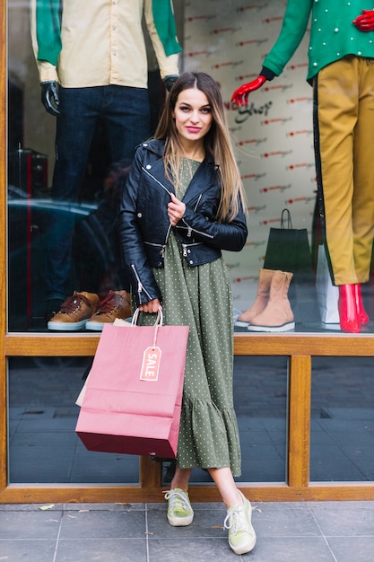 Free photo portrait of a young woman posing in front of window display holding shopping bags in hand