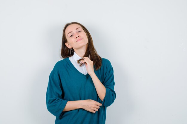 Портрет молодой женщины, позирующей, держась за прядь в свитере над белой рубашкой и гордо смотрящей спереди