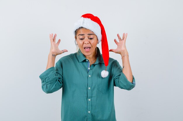 Портрет молодой женщины, поднимающей руки, крича в рубашке, шляпе Санта-Клауса и испуганного вида спереди