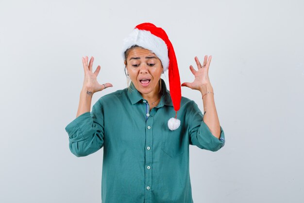 Portrait of young woman raising hands while screaming in shirt, Santa Claus hat and looking horrified front view