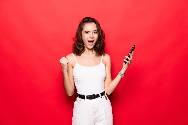 Portrait of young woman screaming and clenching fist like rejoicing victory or triumph isolated over red wall