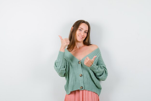 Portrait of young woman showing double thumbs up in cardigan, skirt and looking optimistic front view