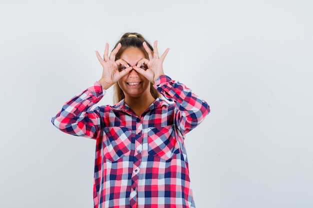 Free photo portrait of young woman showing glasses gesture in casual shirt and looking cheerful front view