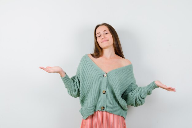 Portrait of young woman showing helpless gesture in cardigan, skirt and looking optimistic front view