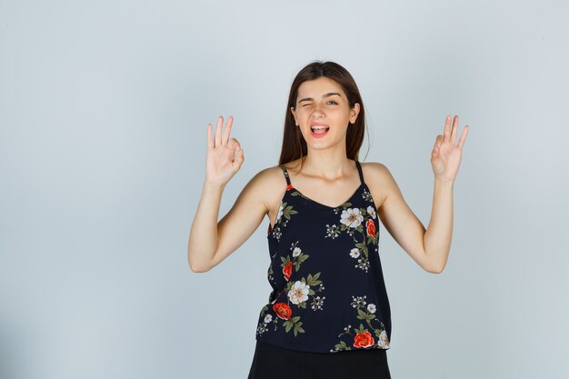 Портрет молодой женщины, показывающей жест ОК, моргая в блузке и глядя на веселый вид спереди