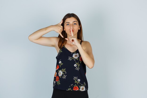 Portrait of young woman showing phone gesture and silence sign in blouse, skirt and looking careful front view