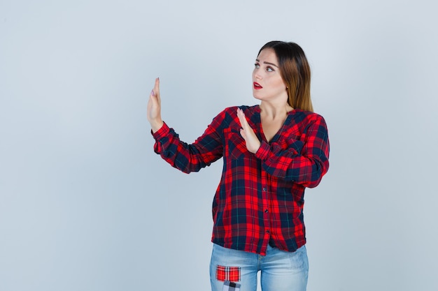 Portrait of young woman showing stop gesture as if trying to defend herself in checked shirt and looking frightened front view