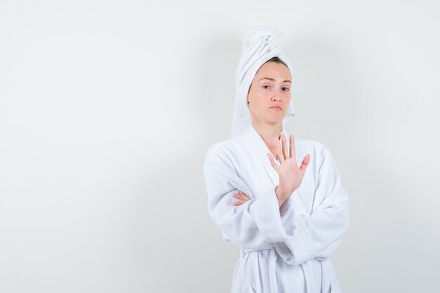 Портрет молодой женщины, показывающей жест стоп в белом халате, полотенце и уверенно выглядящей вид спереди