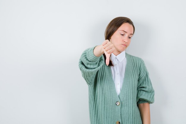 Portrait of young woman showing thumb down, looking down in blouse, cardigan and looking upset front view