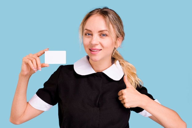 Портрет молодой женщины, показывая большой палец вверх жест, удерживая белый пустой визитная карточка против голубой стене