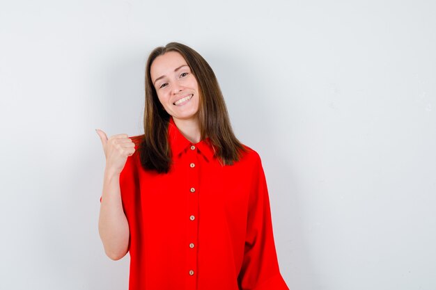 Портрет молодой женщины показывает палец вверх в красной блузке и смотрит веселый вид спереди