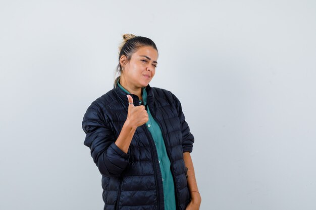 Portrait of young woman showing thumb up in shirt, puffer jacket and looking lucky front view