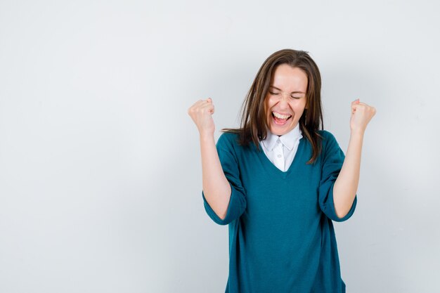 Portrait of young woman showing winner gesture in sweater over white shirt and looking blissful front view