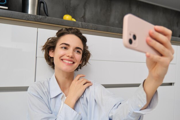 Portrait of young woman sits on kitchen floor with telephone takes selfie on smartphone with app fil