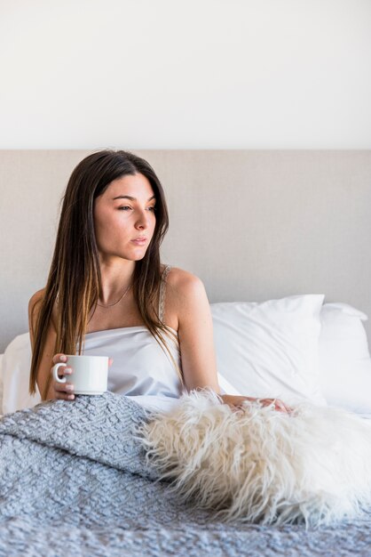 Portrait of a young woman sitting on bed holding coffee cup