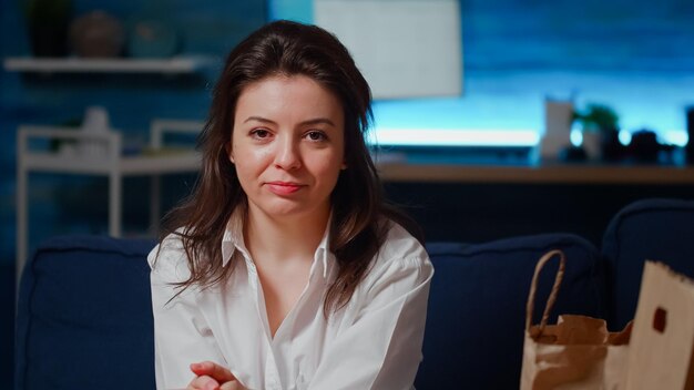 Portrait of young woman sitting in living room eating snack after work. Person with chips and delivery bag on couch looking at camera and smiling. Adult relaxing with takeaway meal