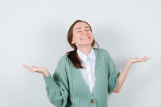 Portrait of young woman spreading palms sideways, keeping eyes shut in blouse, cardigan and looking merry front view