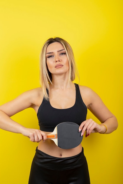 Free photo portrait of young woman standing and holding table tennis racket on yellow wall.