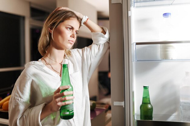 Portrait of young woman standing on kitchen at night and looking in opened fridge while holding beer in hand at home