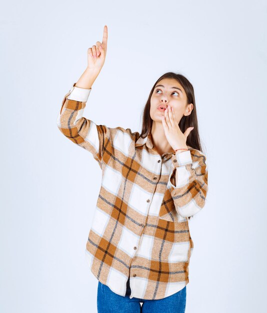 Portrait of young woman standing and pointing at somewhere on white wall. 