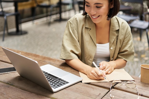 Portrait of young woman studying online sitting with laptop writing down making notes and looking at