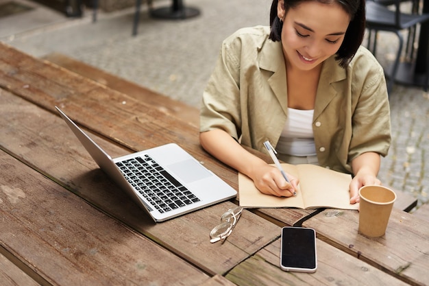 Portrait of young woman studying online sitting with laptop writing down making notes and looking at