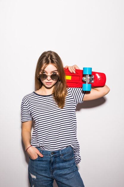 Portrait of a young woman in sunglasses posing with skateboard while standing over white wall