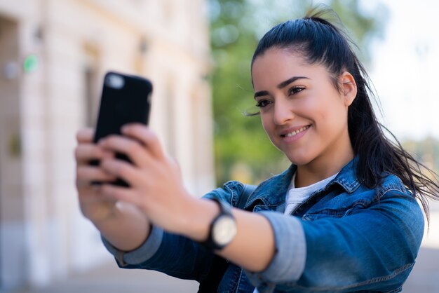 야외에서 서있는 동안 그녀의 mophile 전화로 셀카를 복용하는 젊은 여자의 초상화