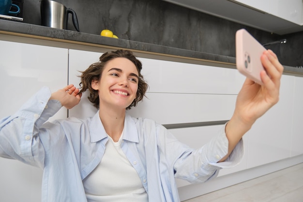 Portrait of young woman video chats records vlog while sits on kitchen floor cute girl takes selfie