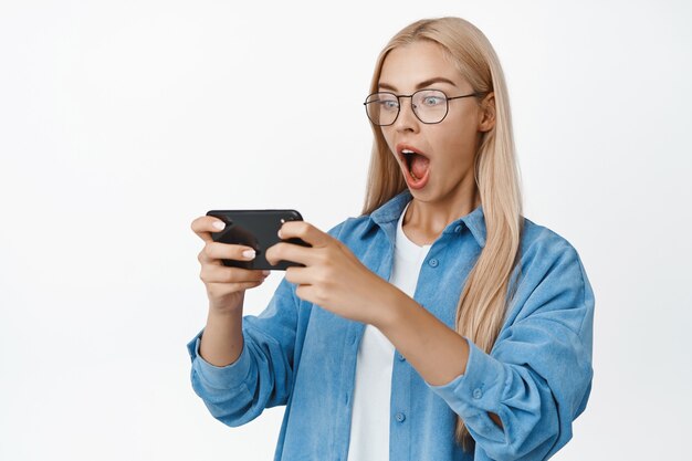 Portrait of young woman watching video on smartphone with shocked face expression. Girl widen eyes and stare at mobile phone, standing on white.