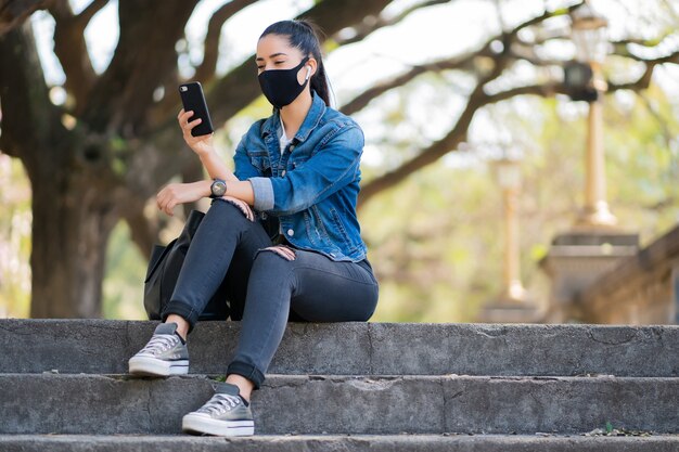 フェイスマスクを着用し、屋外の階段に座っている間彼女の携帯電話を使用して若い女性の肖像画