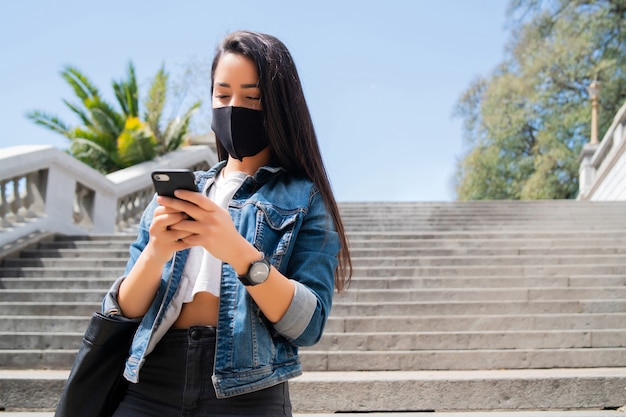 Portrait of young woman wearing protective mask and using her mobile phone while standing outdoors on the street. New normal lifestyle concept. Urban concept.