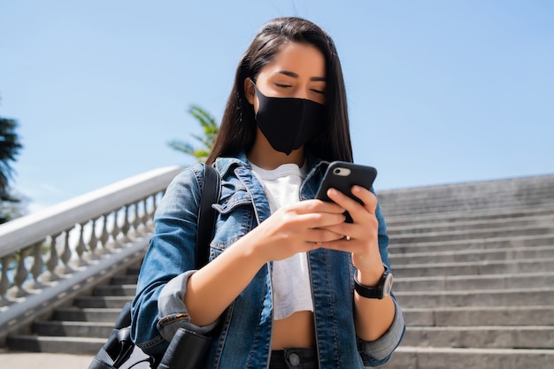 Портрет молодой женщины в защитной маске и с помощью своего мобильного телефона, стоя на открытом воздухе на улице.