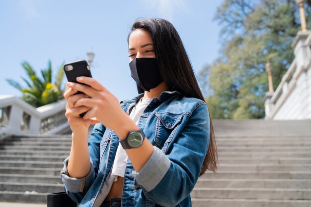 Портрет молодой женщины в защитной маске и с помощью своего мобильного телефона, стоя на открытом воздухе на улице.