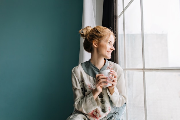 大きな窓の横にあるコーヒーやお茶を飲んで、笑って、自宅で幸せな朝を楽しんでいるブロンドの髪を持つ若い女性の肖像画。ターコイズブルーの壁。花にシルクのパジャマを着ています。