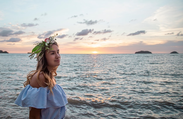 Portrait of a young woman with a composition of flowers on her head against the background of the ocean at sunset.