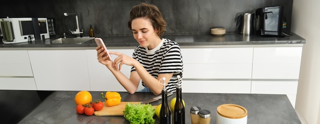 Free photo portrait of young woman with smartphone sitting in the kitchen and cooking salad searching for