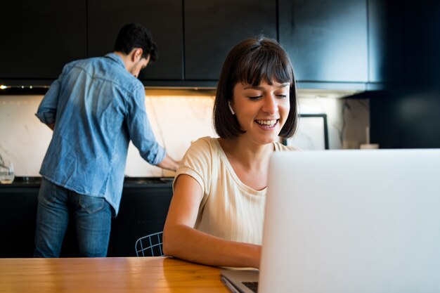 Portrait of young woman working with a laptop from home while man cleaning kitchen at background.