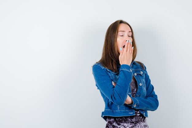 Portrait of young woman yawning while keeping hand on mouth in denim jacket, dress and looking drowsy front view