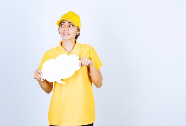 Portrait of a young woman in yellow uniform holding an empty white speech bubble cloud . 