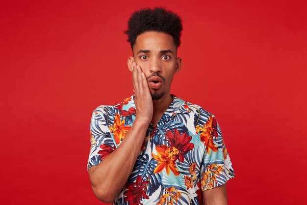 Portrait of young wondered african american guy, wears in hawaiian shirt, looks at the camera with surprised expression touches cheek, stands over red background