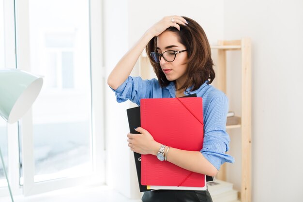 A portrate of brunnete girl in blue shirt standing in office. She holds folders and looks disappointed.