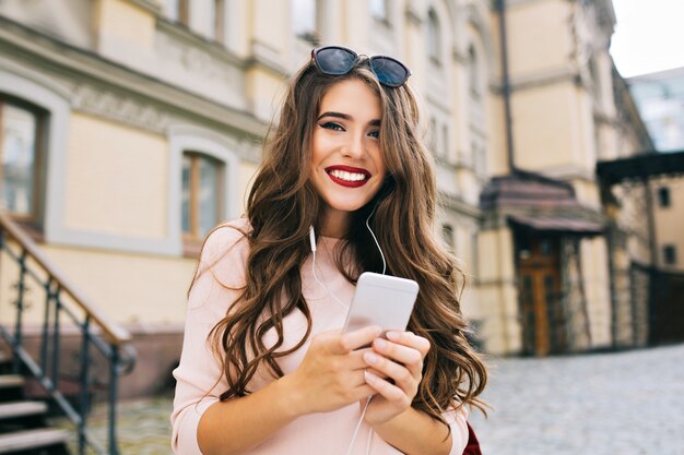 Портрет милой девушки с длинными вьющимися волосами и телефоном в руках, улыбаясь в городе на фоне здания